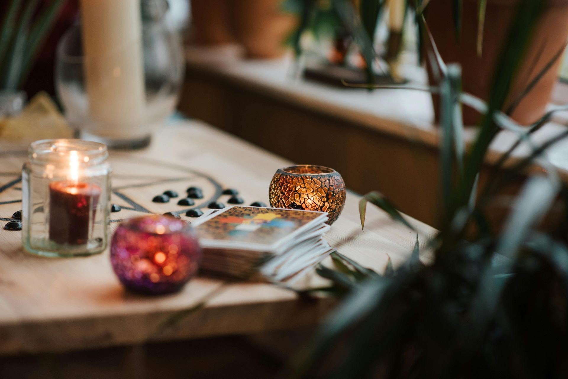 Las velas iluminan las cartas del tarot sobre una mesa de madera, con plantas y una ventana al fondo.