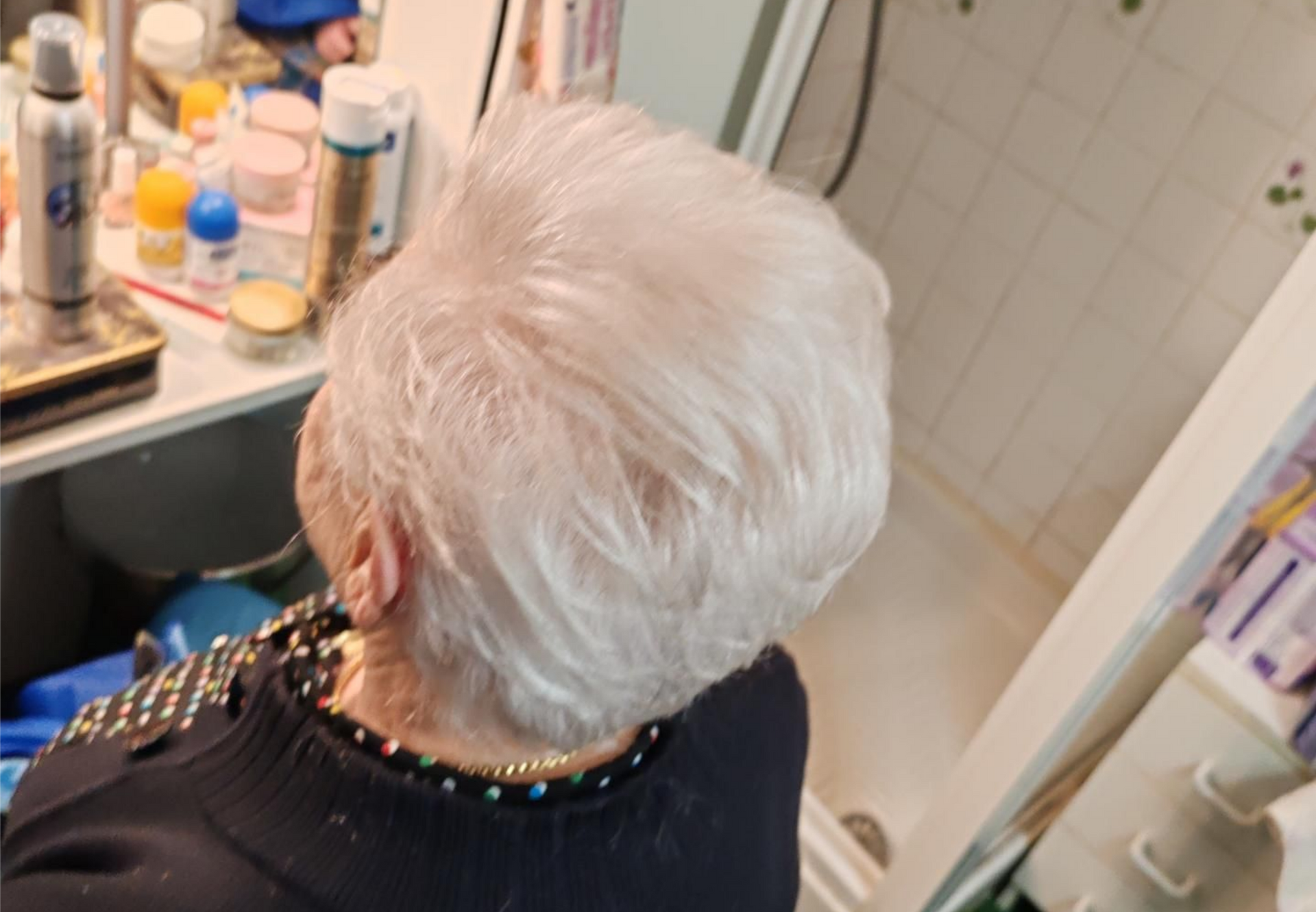 Une personne aux cheveux courts et blancs, dans une salle de bains devant un miroir.