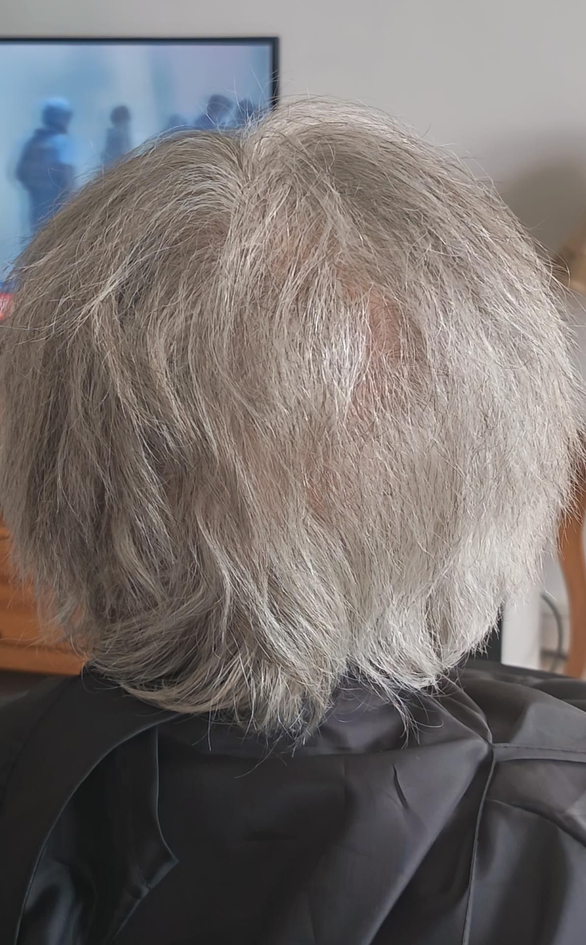 Vue de dos d'une personne aux cheveux gris non coiffés, portant une blouse noire.