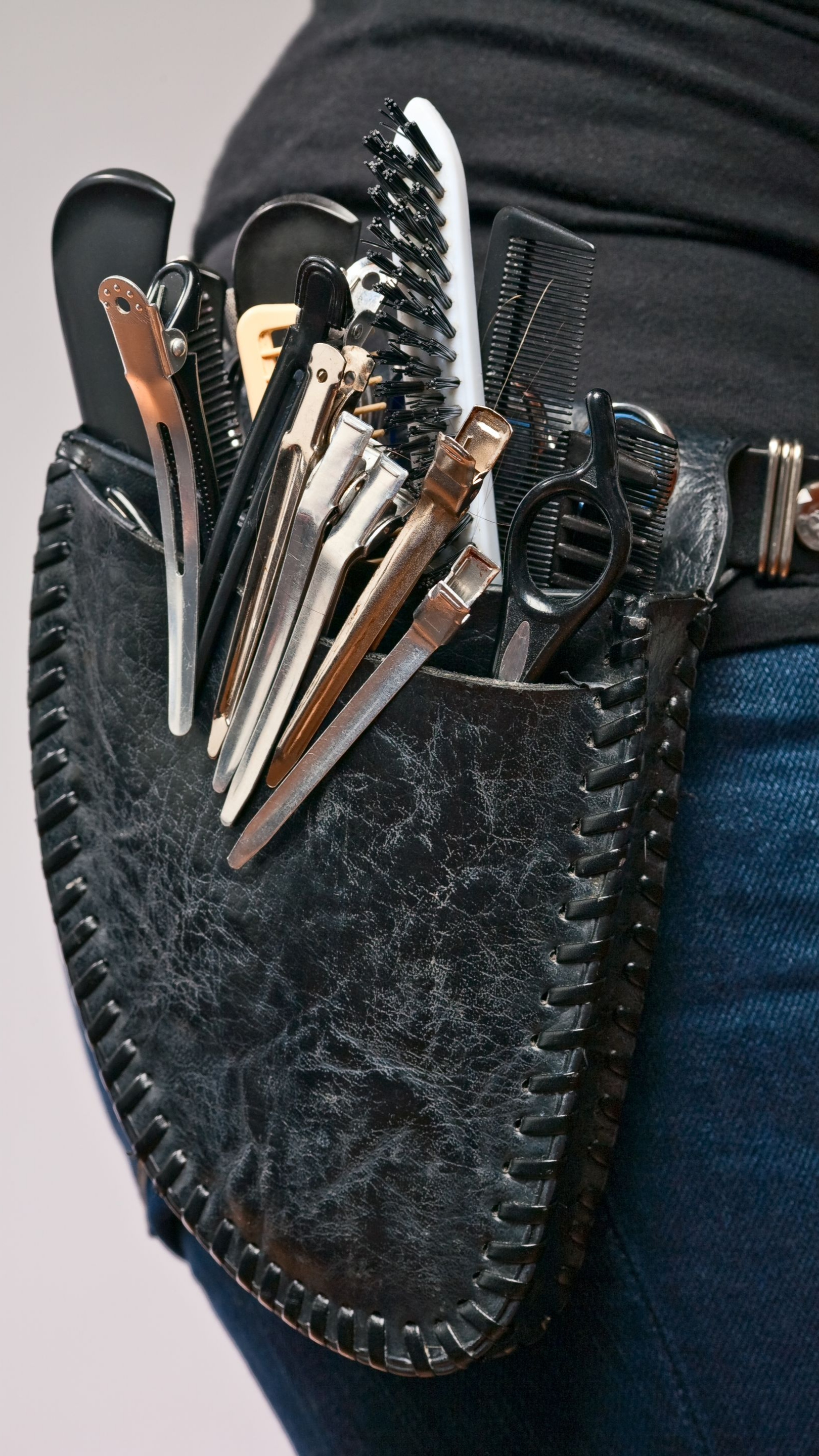 Ceinture à outils de coiffeur en cuir noir avec divers peignes, pinces et ciseaux, portée à la hanche.