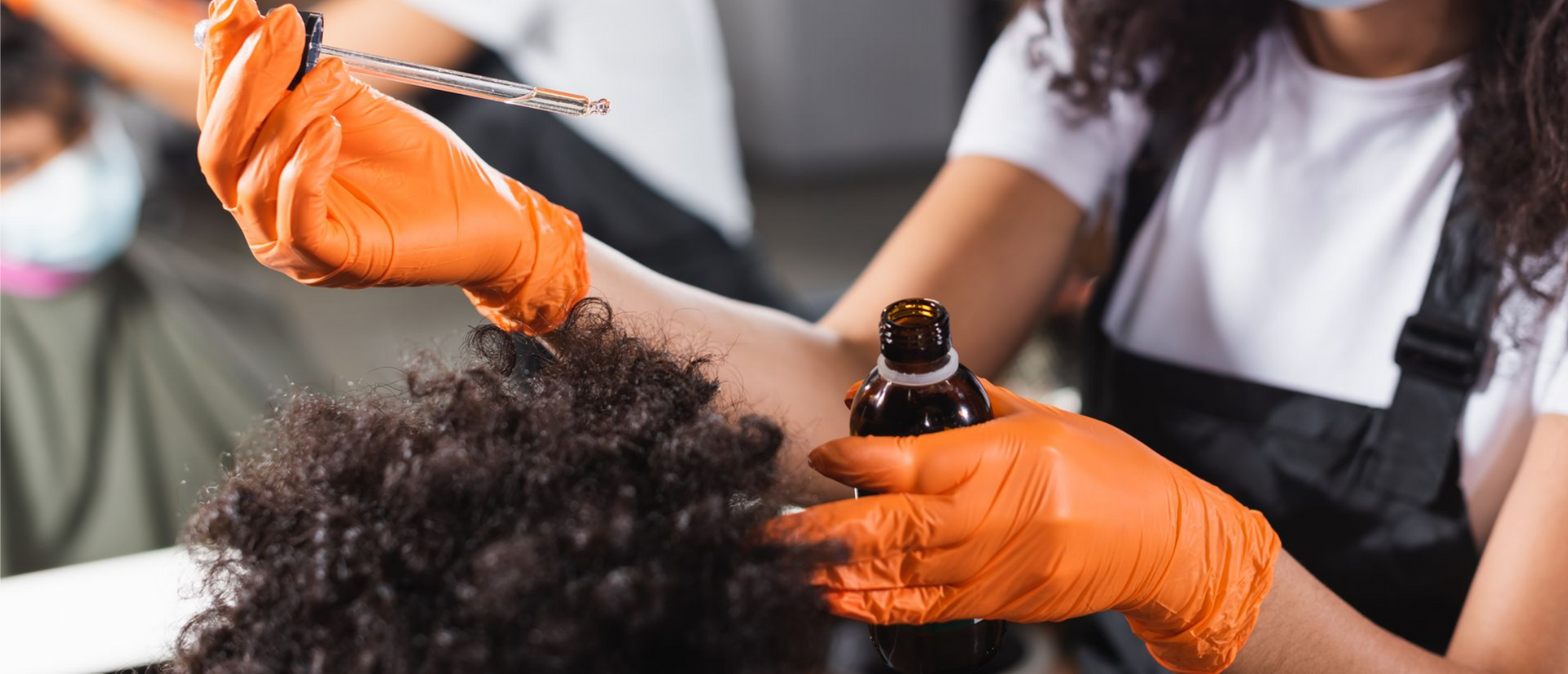 Une personne applique un sérum sur des cheveux bouclés dans un salon de coiffure, portant des gants orange.