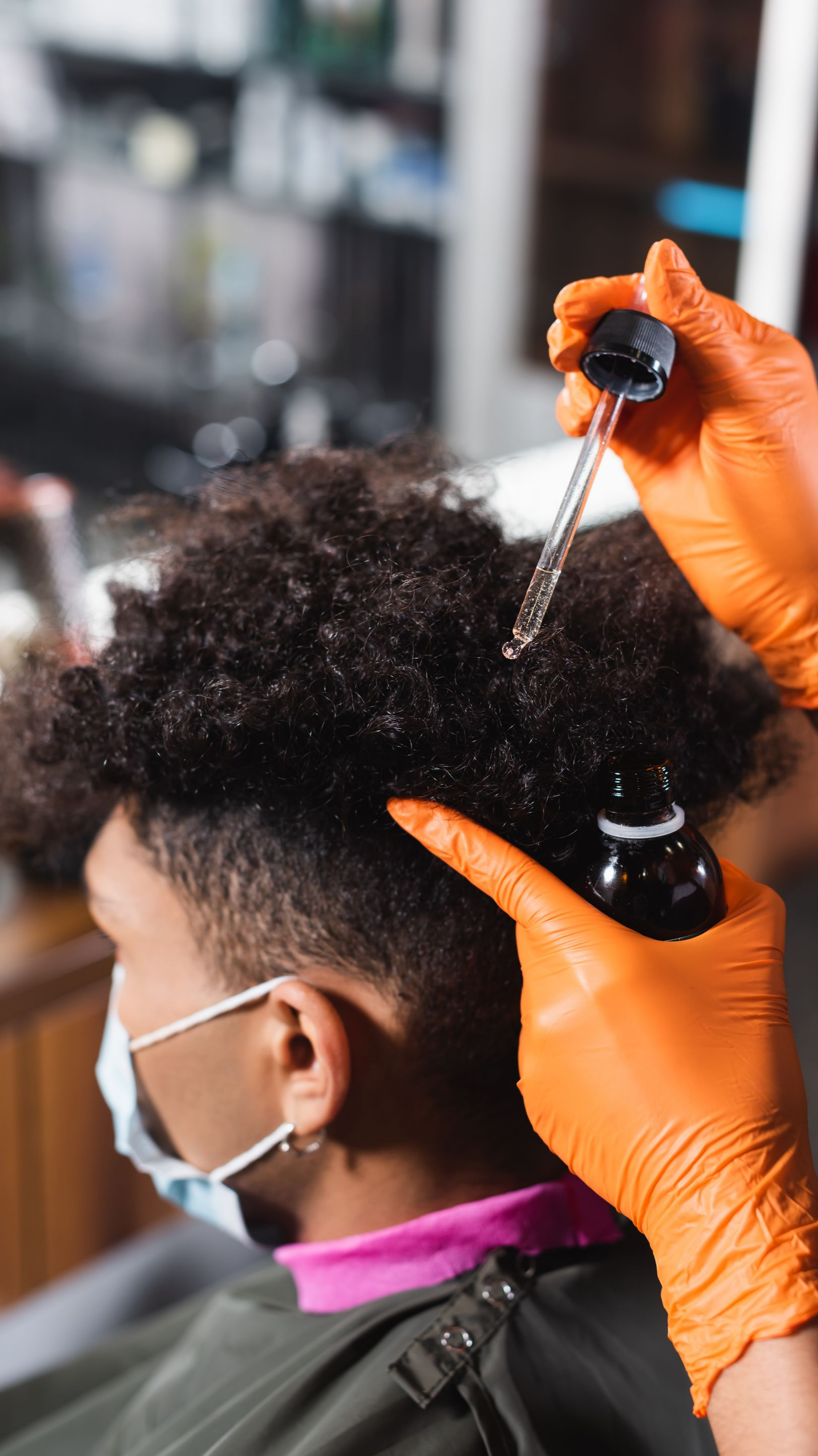 Une personne aux cheveux bouclés, portant un masque, se fait appliquer de l'huile dans un salon de coiffure.