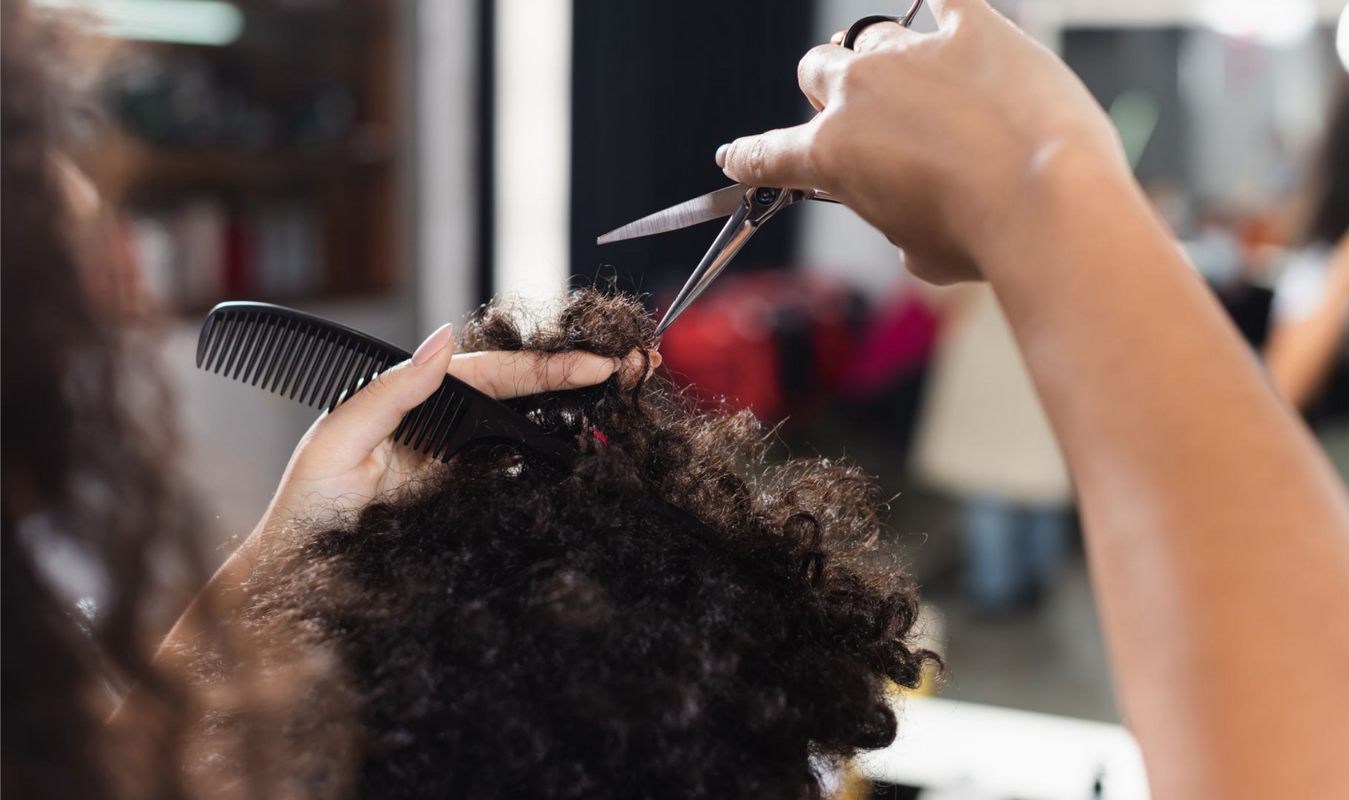 Des mains utilisent des ciseaux et un peigne pour couper des cheveux bouclés dans un salon de coiffure.