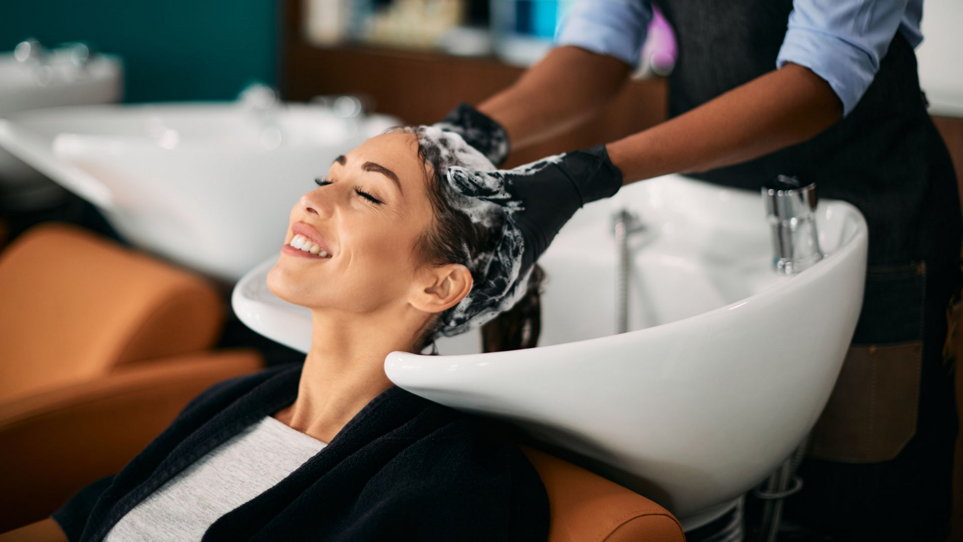 Une femme sourit pendant qu'on lui lave les cheveux dans le lavabo du salon.