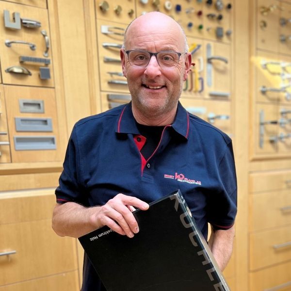 Ein kahlköpfiger Mann mit Brille und blauem Hemd hält eine schwarze Schachtel mit der Aufschrift „kdc“ darauf.