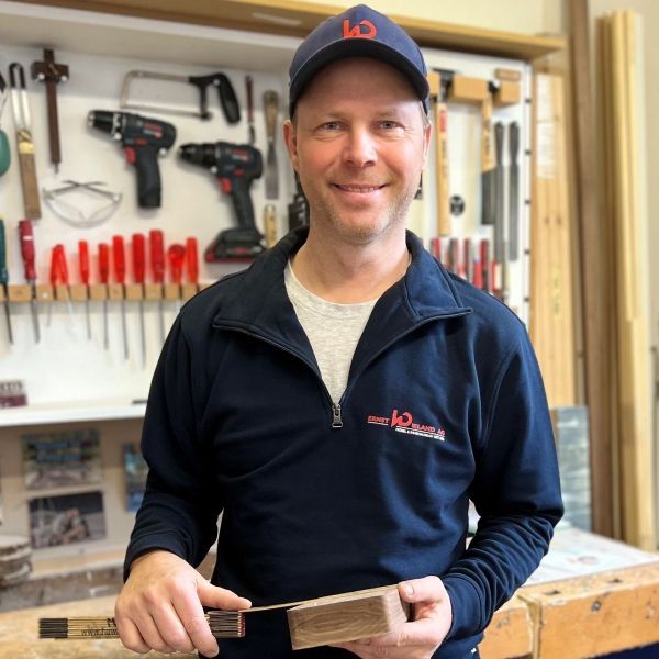 Ein Mann hält ein Stück Holz vor einer Wand voller Werkzeuge