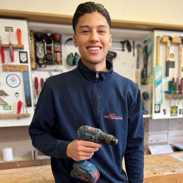 Ein Mann in einem blauen Sweatshirt hält eine Bohrmaschine und lächelt