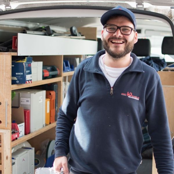 Ein Mann mit Brille und Hut lächelt vor einem Lieferwagen