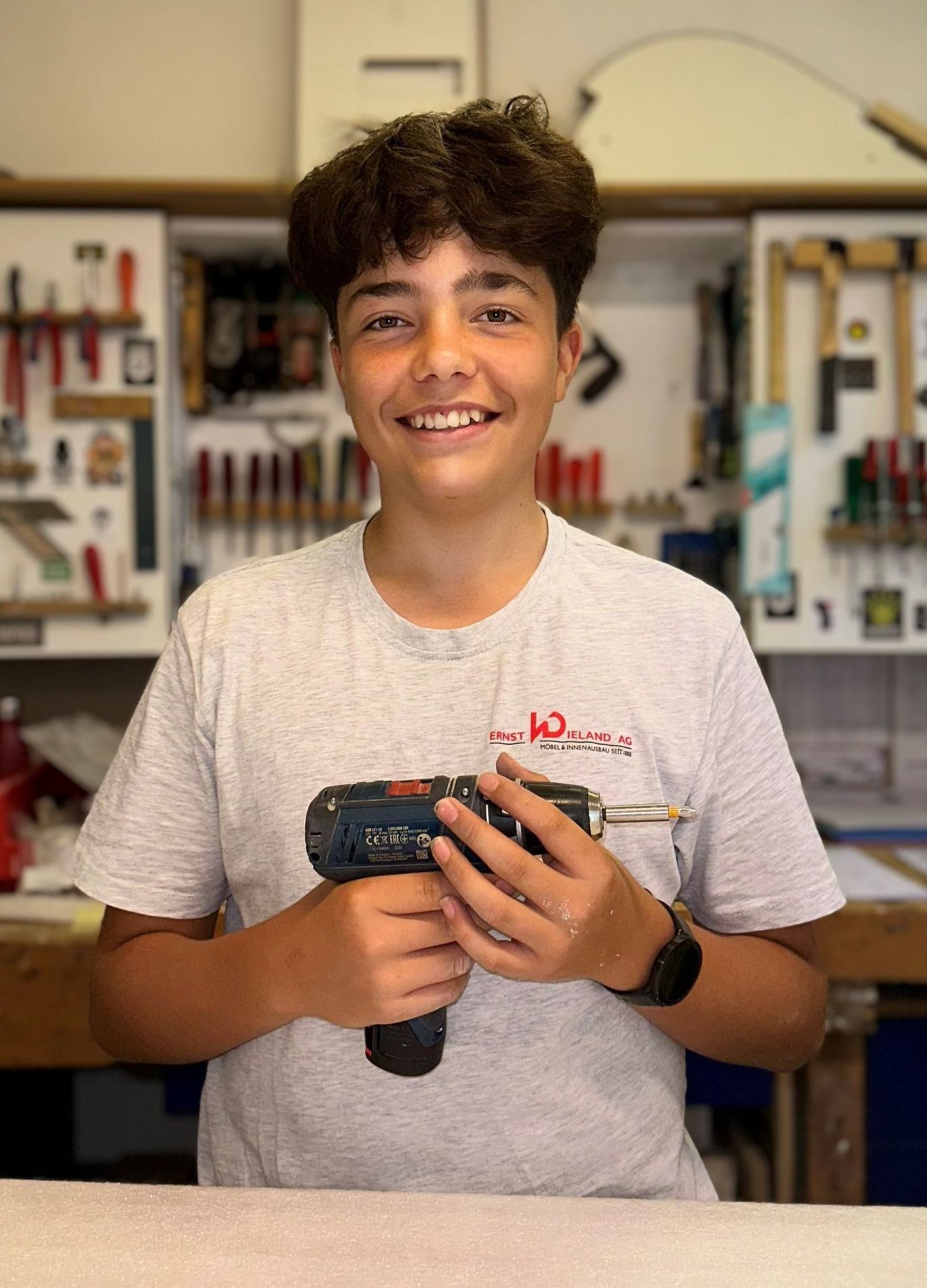 Ein kleiner Junge hält in einer Werkstatt eine Bohrmaschine.