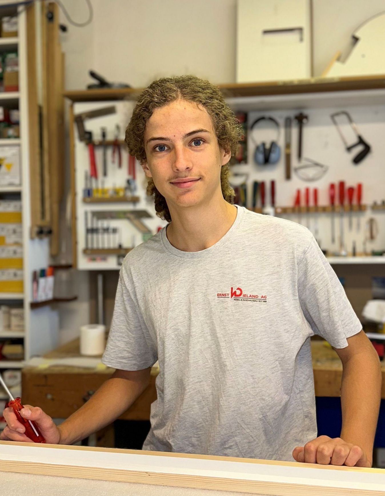 Ein junger Mann in einem grauen T-Shirt steht vor einem Holztisch.