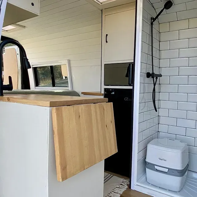 Interior de una furgoneta camper, con ducha, inodoro, zona de cocina con mesa plegable y armarios.