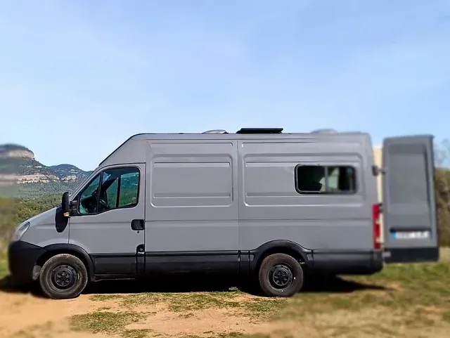 Autocaravana gris estacionada al aire libre, puerta trasera abierta, fondo de montañas.