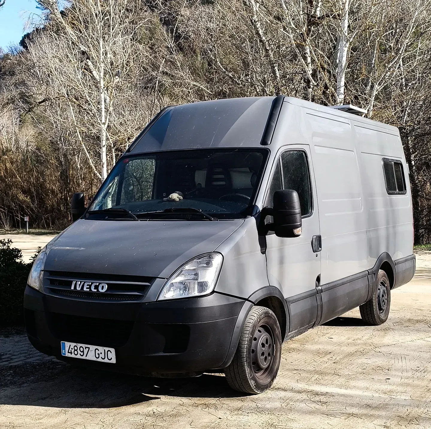 Autocaravana gris estacionada al aire libre. Árboles al fondo.