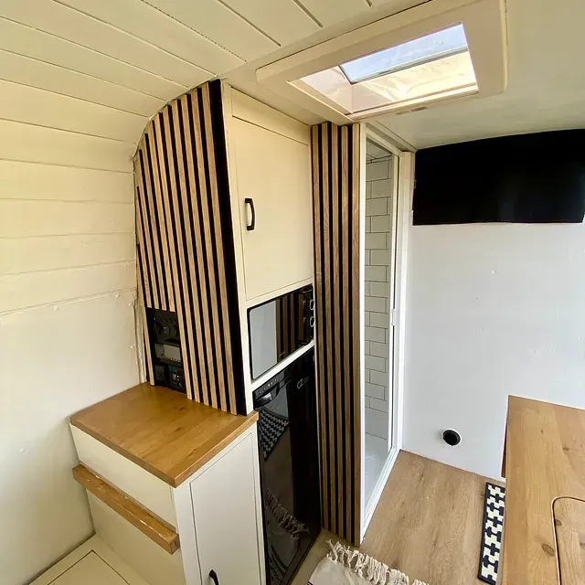 Interior de una autocaravana con cocina, ducha y detalles en madera. Paredes blancas, suelo color canela y claraboya.