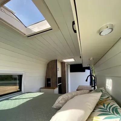 Interior de una camioneta, que muestra una cama, un tragaluz, un área de cocina y paredes de madera blanca.
