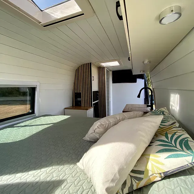Interior de una caravana con cama, almohadas, detalles de madera y tragaluces.