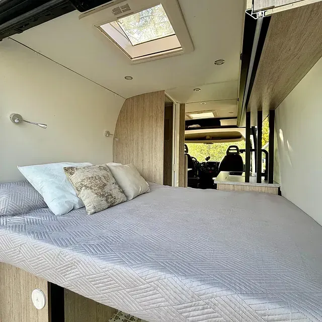 Interior de una furgoneta camper, con una cama con almohadas, tragaluz y vista del asiento del conductor.