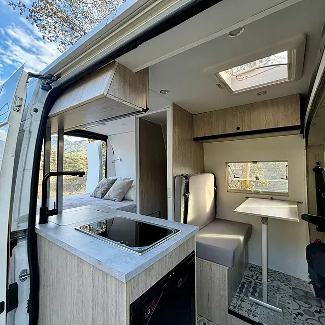 Interior de una autocaravana con cocina, cama y comedor. Armarios de madera clara y detalles en gris.