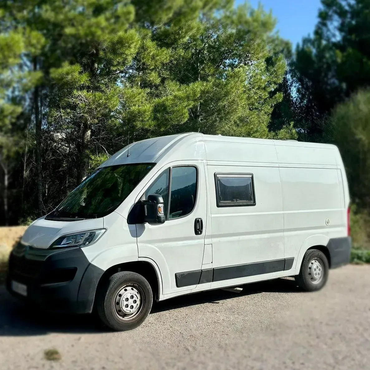 Autocaravana blanca estacionada en un camino de grava con árboles al fondo.