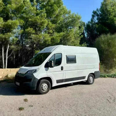 Autocaravana blanca estacionada en una zona pavimentada, árboles al fondo, día soleado.