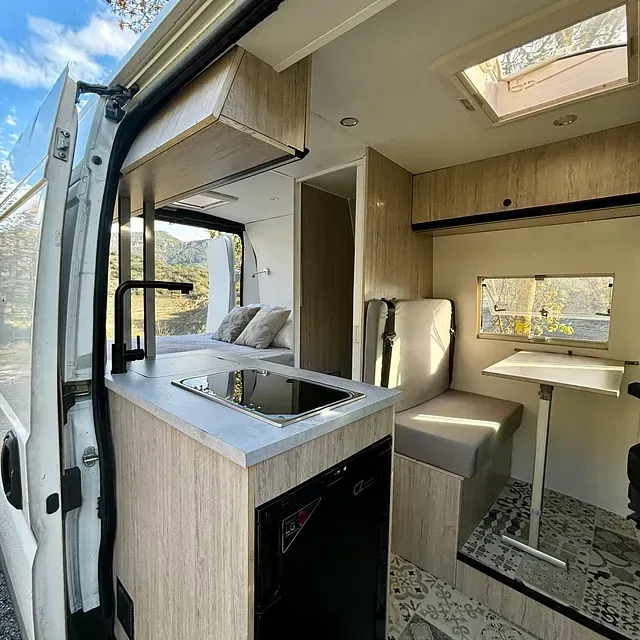Interior de una autocaravana, con cocina, zona de estar y cama, todo ello con paneles de madera clara.
