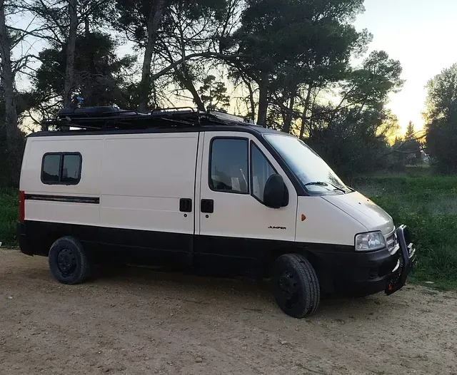 Furgoneta camper de color crema y negro, con portaequipaje, estacionada en un camino de tierra con árboles al fondo.