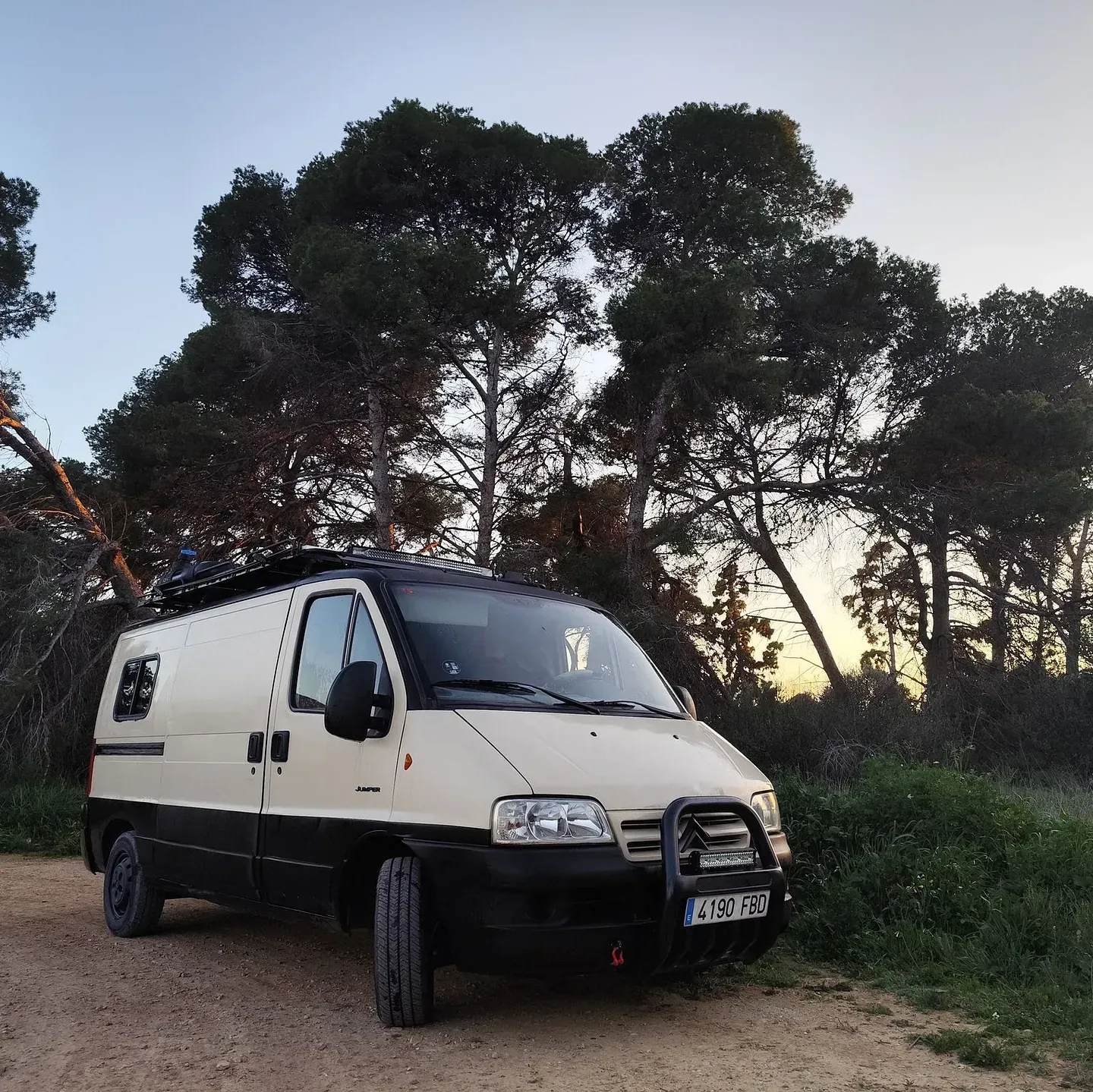 Autocaravana de color tostado y negro estacionada en un camino de tierra frente a una zona boscosa al anochecer.