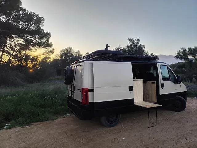 Autocaravana blanca con fondo negro aparcada al aire libre al atardecer, con mesa extraíble.