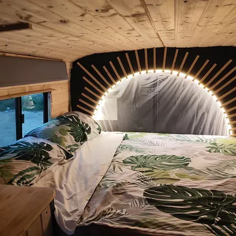 Acogedor dormitorio tipo furgoneta con paneles de madera, ropa de cama con estampado tropical y cabecera retroiluminada.