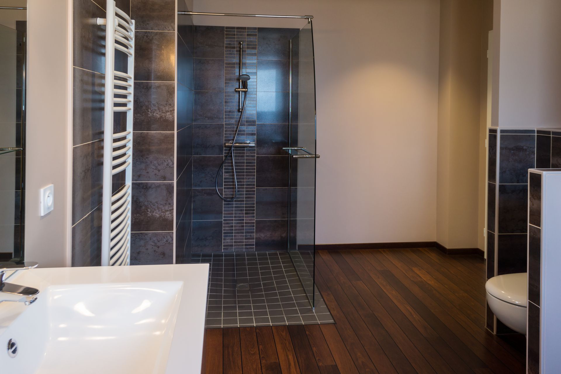 Salle de bains moderne avec douche vitrée, carrelage foncé, parquet et meuble-lavabo blanc.