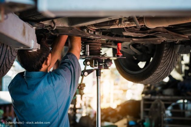 In einer Autowerkstatt arbeitet ein Mechaniker am Unterboden eines auf einer Hebebühne angehobenen Autos.
