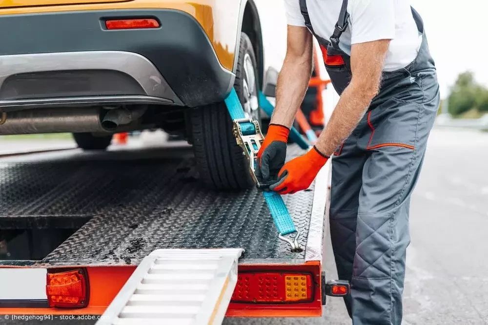 Ein Arbeiter mit orangefarbenen Handschuhen sichert ein gelbes Auto mit blauen Spanngurten auf einem Tieflader.