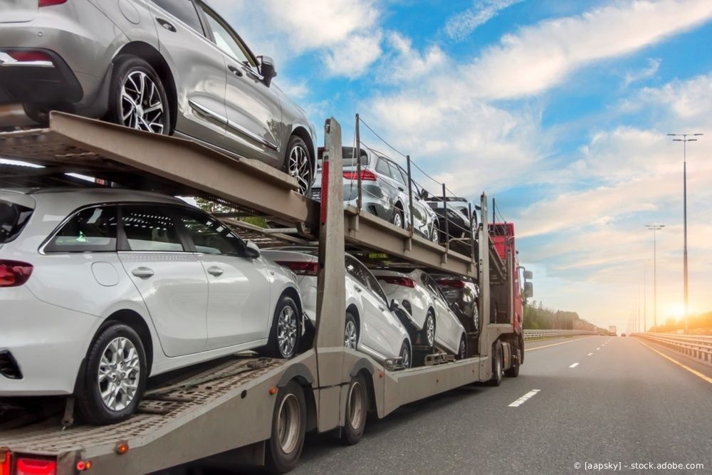 Ein mit mehreren neuen weißen und silbernen SUVs beladener Autotransporter fährt bei Sonnenuntergang auf einer Autobahn.