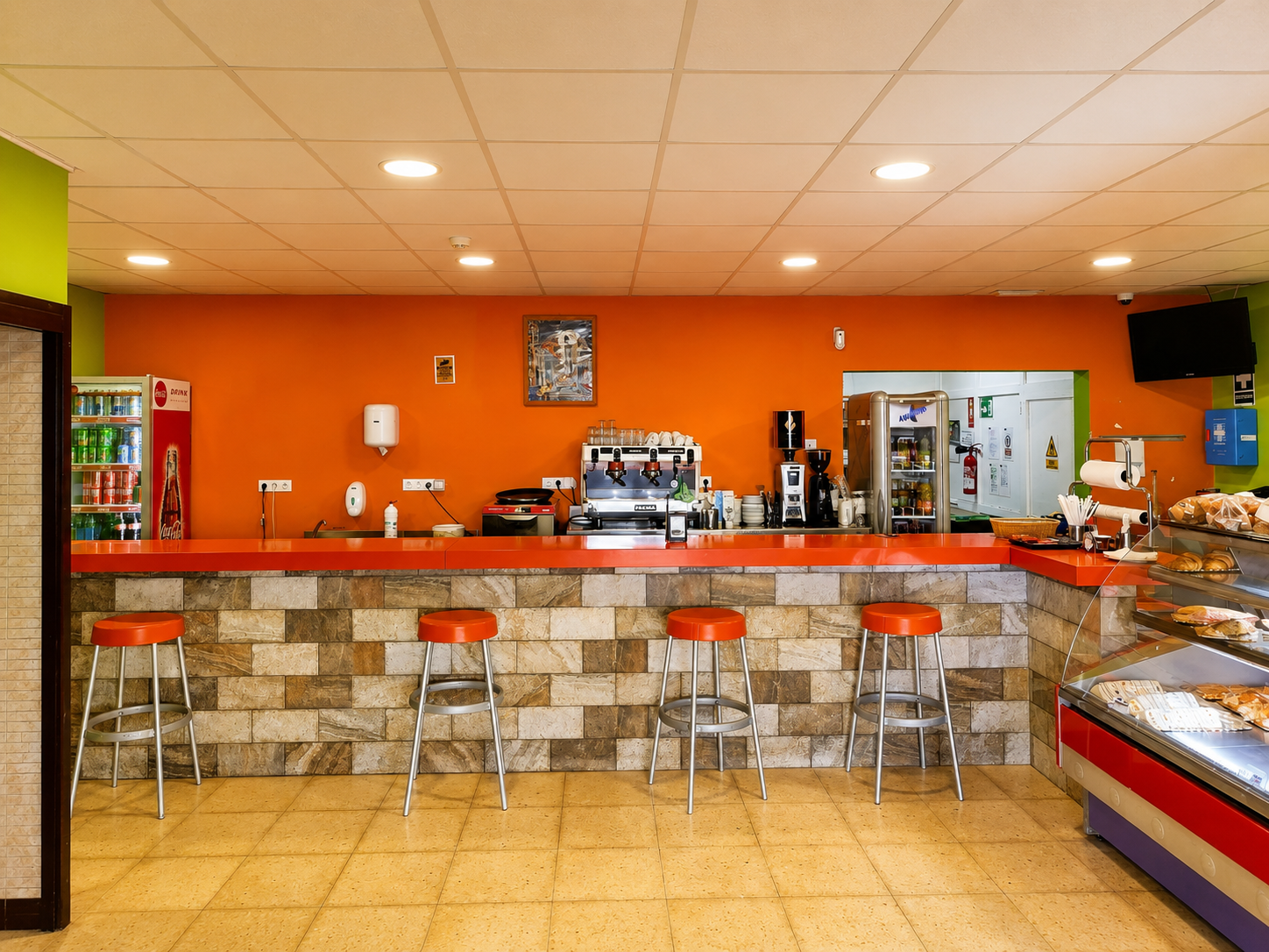 Mostrador de cafetería naranja vacío con taburetes, máquina de café y vitrina refrigerada dentro de un interior luminoso con azulejos.