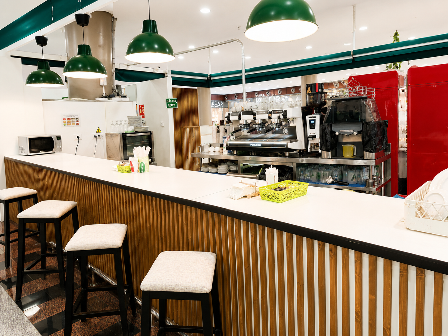 Interior luminoso de una cafetería con una larga barra blanca, lámparas colgantes verdes y taburetes de bar.