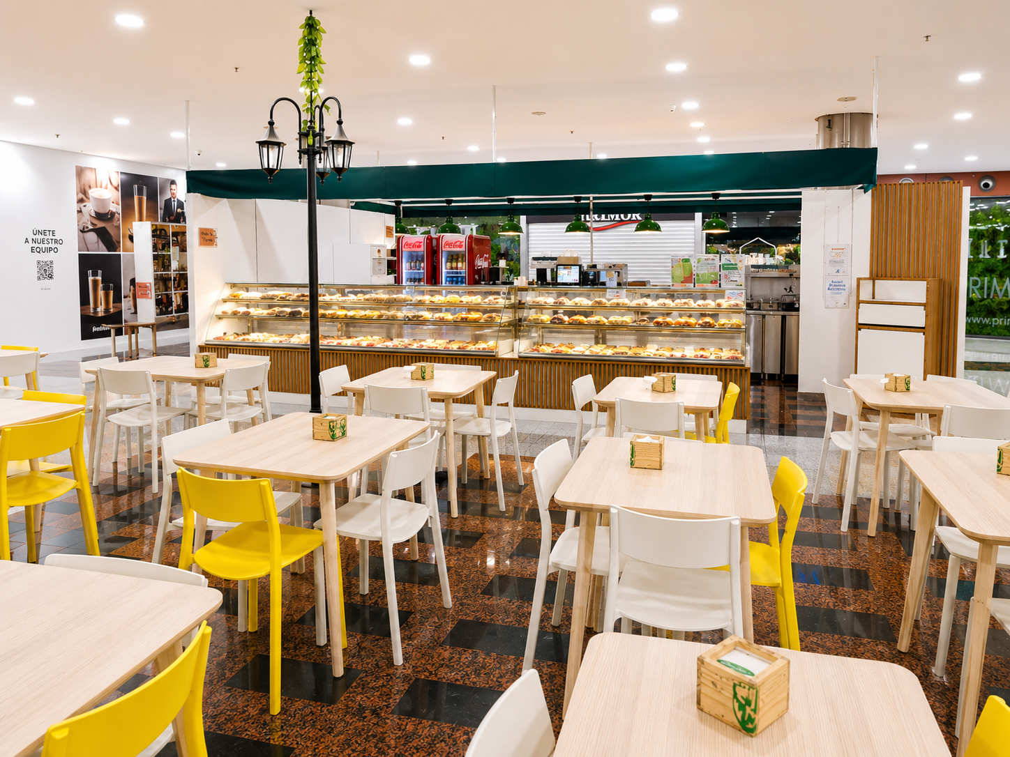 Cafetería luminosa con mesas blancas, sillas amarillas y un largo mostrador de comida bajo una decoración verde.