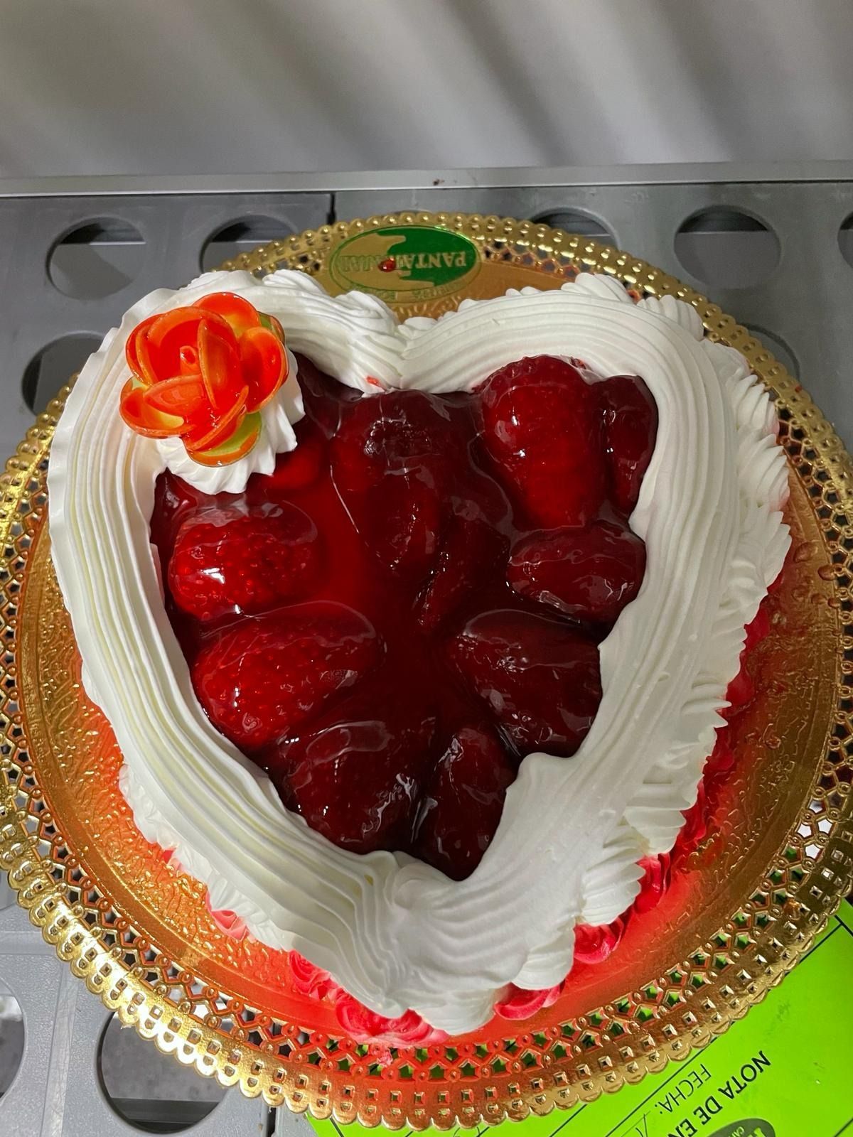 Tarta de fresa con forma de corazón, glaseado blanco y decoración de una rosa roja sobre una bandeja dorada.