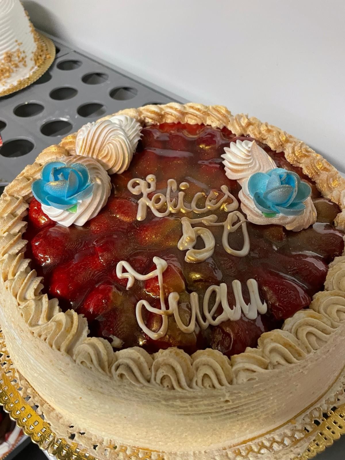 Pastel de chocolate con cobertura roja, flores de glaseado azul y la inscripción blanca “Feliz…” en una mesa.