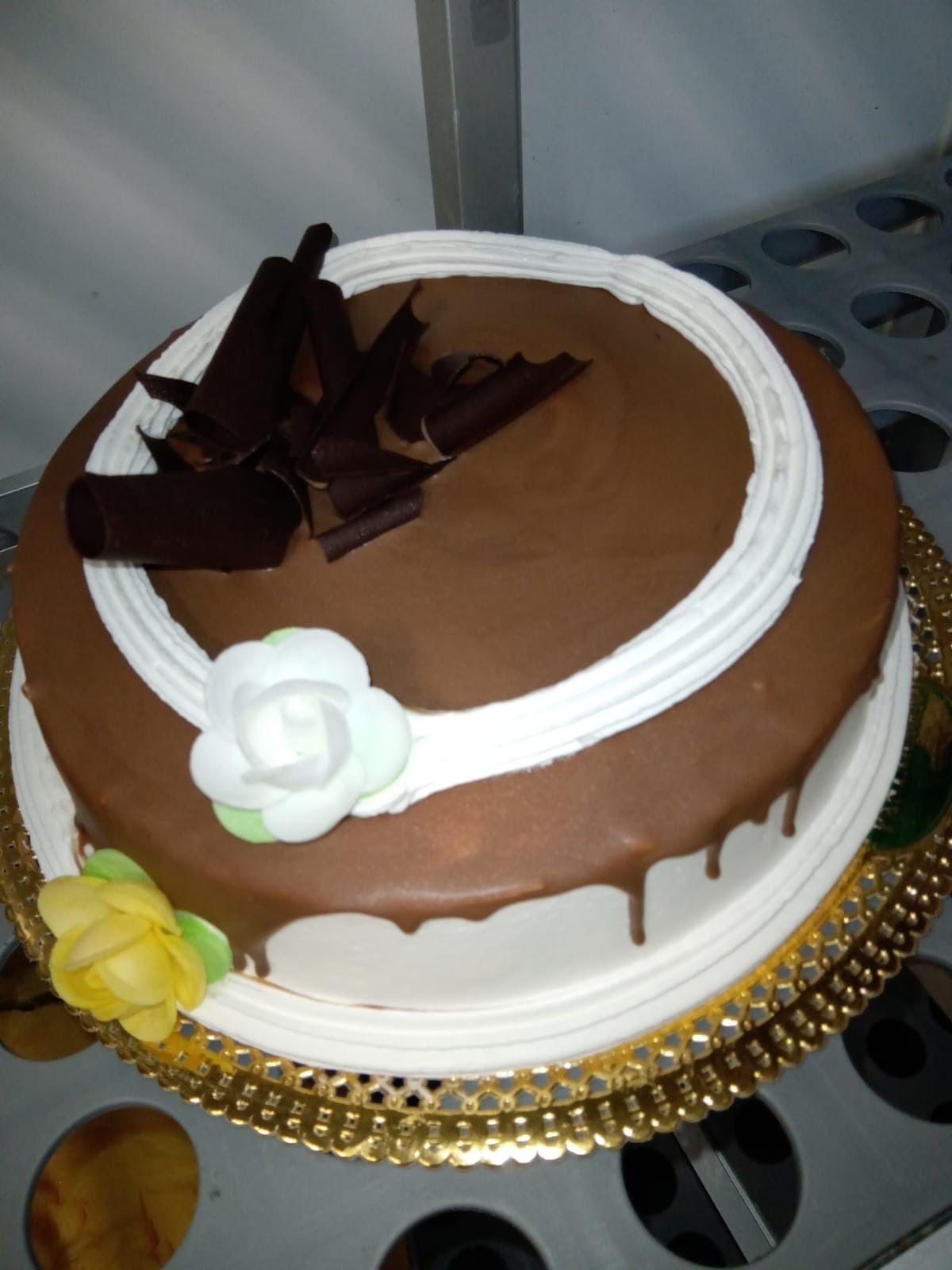 Pastel redondo glaseado de chocolate con decoraciones blancas y virutas de chocolate sobre una bandeja dorada.
