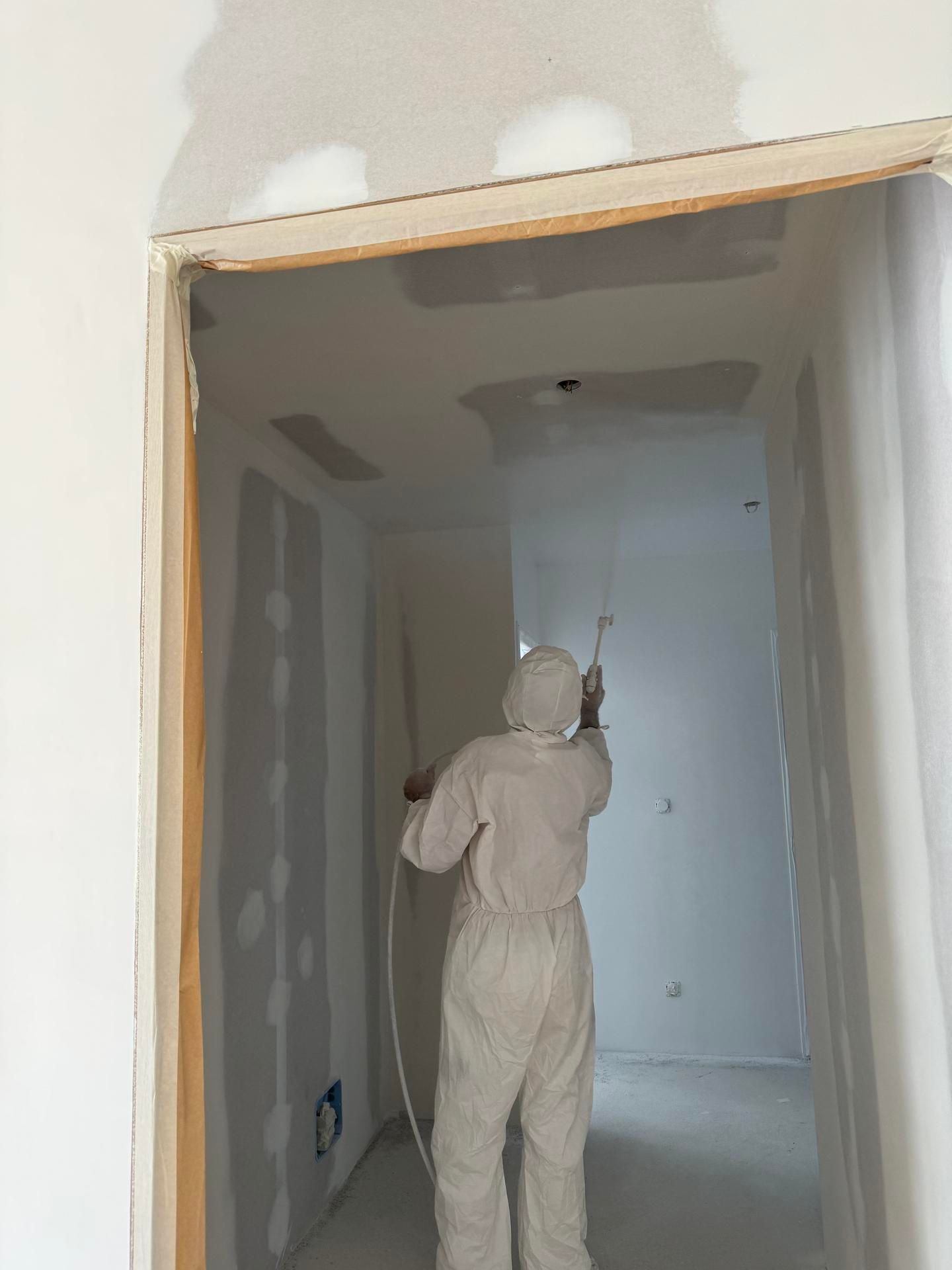 Une personne peint une cloison sèche dans l'encadrement d'une porte de couloir avec un rouleau, vêtue de vêtements de travail blancs et d'une casquette.