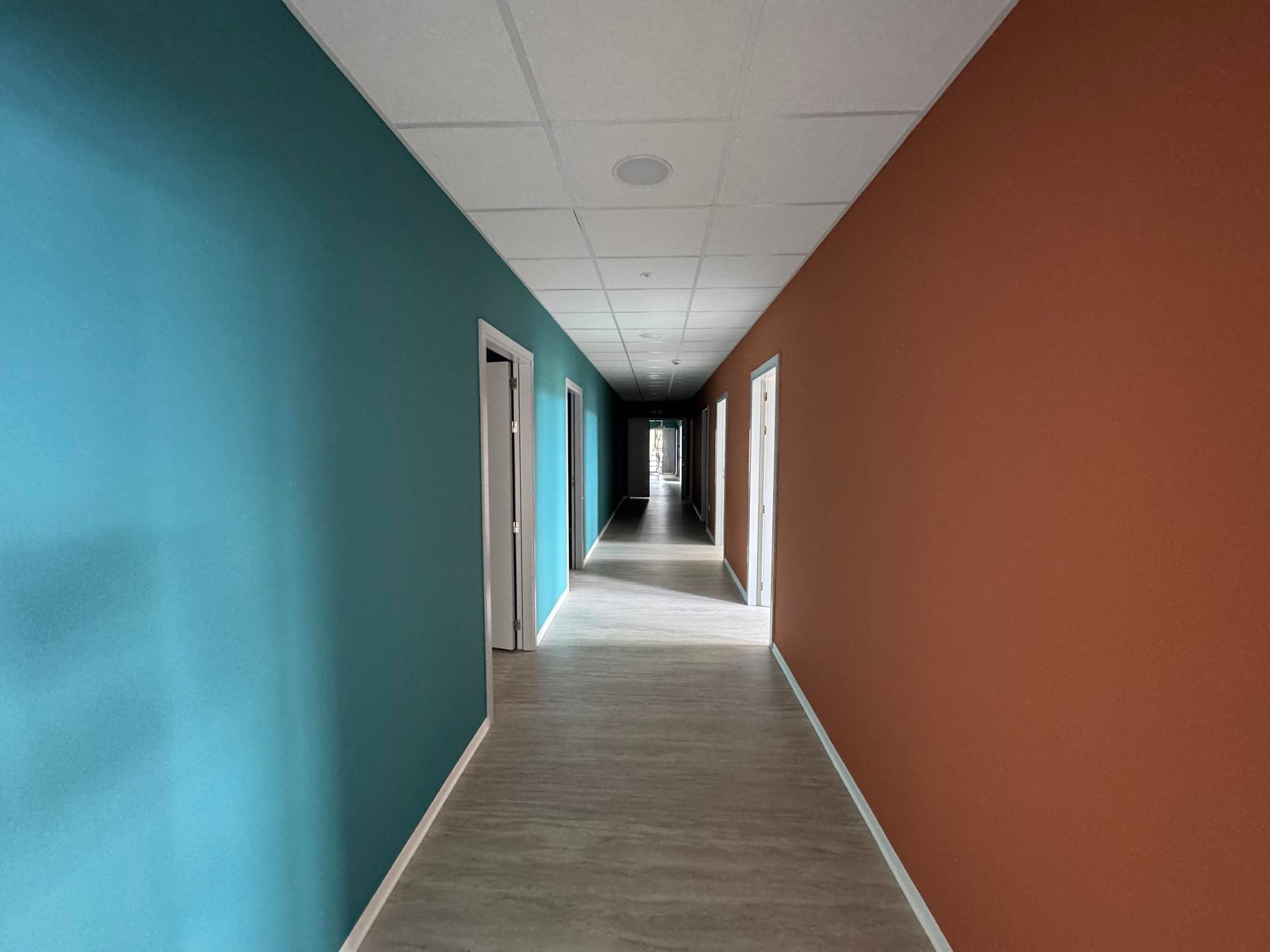 Couloir vide aux murs turquoise et rouille, plafond blanc et long parquet clair menant à un fond sombre.