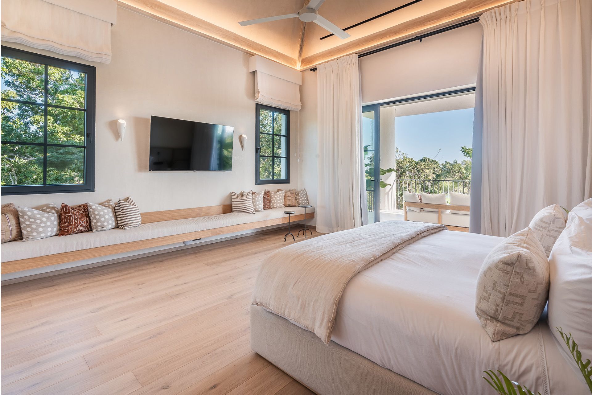 Bright modern bedroom with a wall-mounted TV, window bench, and open doors to a sunny balcony