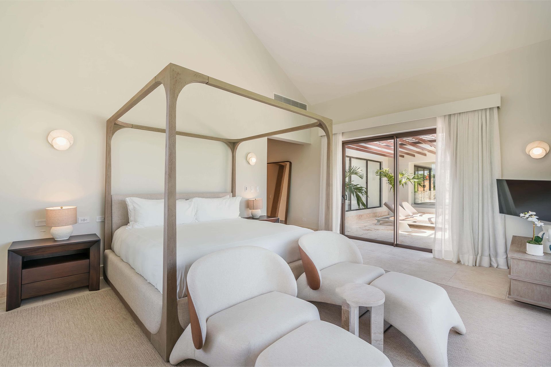 Bright bedroom with a four-poster bed, white chairs, and sliding doors to a patio and pool.