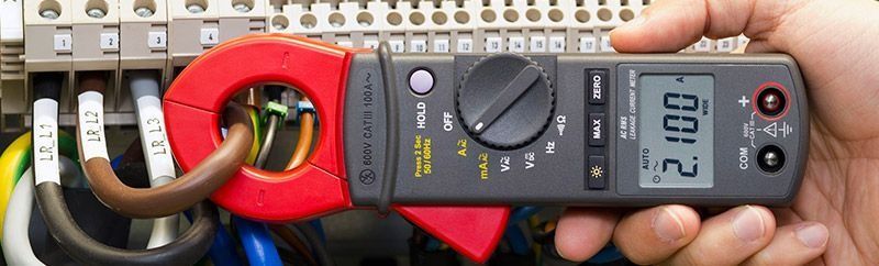 Una mano sostiene un amperímetro de pinza alrededor de cables eléctricos, marcando 2.100.