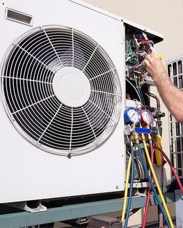 Técnico de climatización realizando el mantenimiento de una unidad exterior de aire acondicionado. Se utilizan manómetros, con los cables a la vista, la carcasa blanca y el ventilador visible.