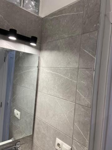 Pared de baño moderna con azulejos grises, espejo con luces y dos enchufes eléctricos.
