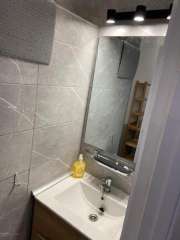 Baño pequeño con azulejos, lavabo blanco, espejo, aplique de pared, dispensador de jabón y estante de madera tipo escalera.