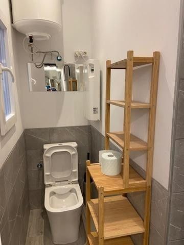 Baño accesible con inodoro elevado, barra de apoyo, papel higiénico y estante de madera junto al inodoro.