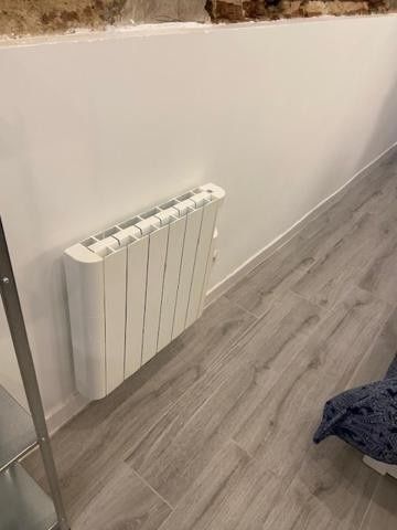 Radiador blanco montado en la pared en una habitación con suelo de madera gris y parte de una tela oscura en el suelo.