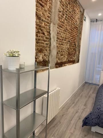 Habitación estrecha con pared de ladrillo visto, estante de cristal, radiador y cortinas transparentes junto a la cama.